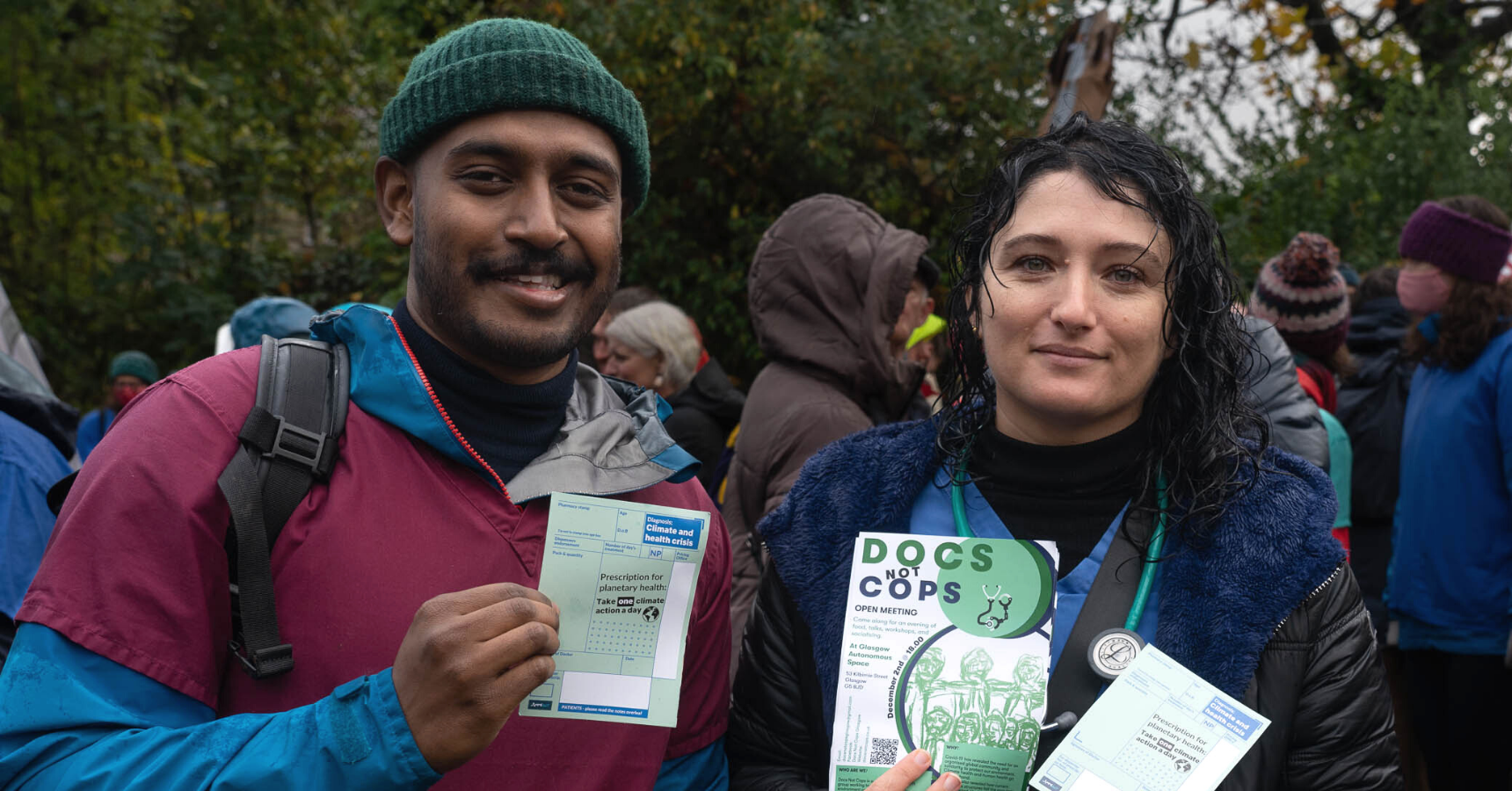 Two people wearing scrubs at a protest holding up pamphlets. The person on the left has brown skin and a green beanie hat, holding a 'prescription for planetary health' pamphlet that looks like a prescription slip. The person on the right holds a 'Docs Not Cops' flyer. They are outside surrounded by people and greenery.