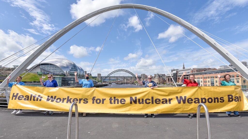 Six health workers on a sunny day on the Millennium Bridge, Newcastle, holding up their arm right arms while holding a large yellow banner with black text reading 'Health Workers for the Nuclear Weapons Ban'. They are looking to the camera and smiling.