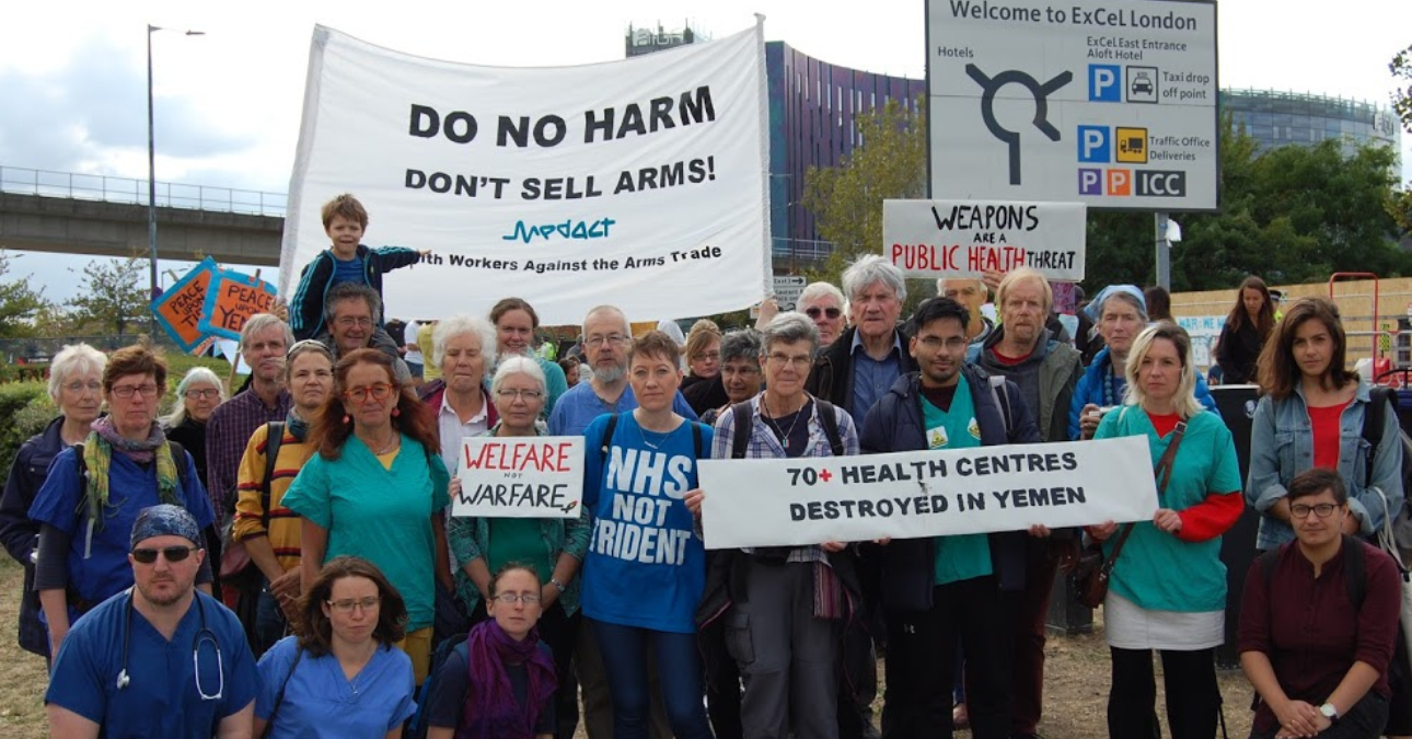 Health workers in scrubs protesting at the Excel Centre in London, with banners including 'Do no harm, Don't sell arms' and '70+ health centres destroyed in Yemen'