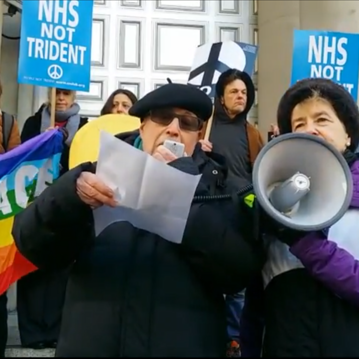People at an NHS Not Trident protest – Michael is doing a speech, reading from a piece of paper into a megaphone - he is wearing a dark coat, sunglasses and a beret