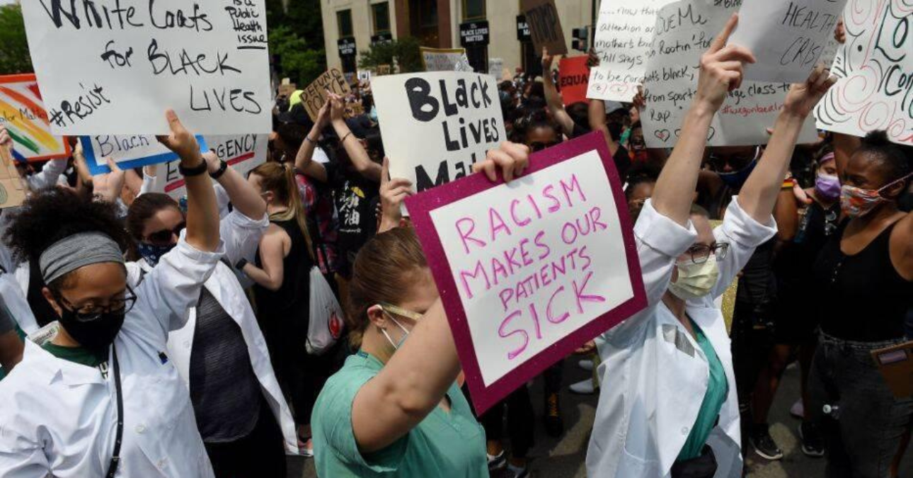 A crowd of protestors, including Black and white people, some wearing scrubs or lab coats, and masks, holding placards such as 'Racism makes us sick' and 'Black Lives Matter'.