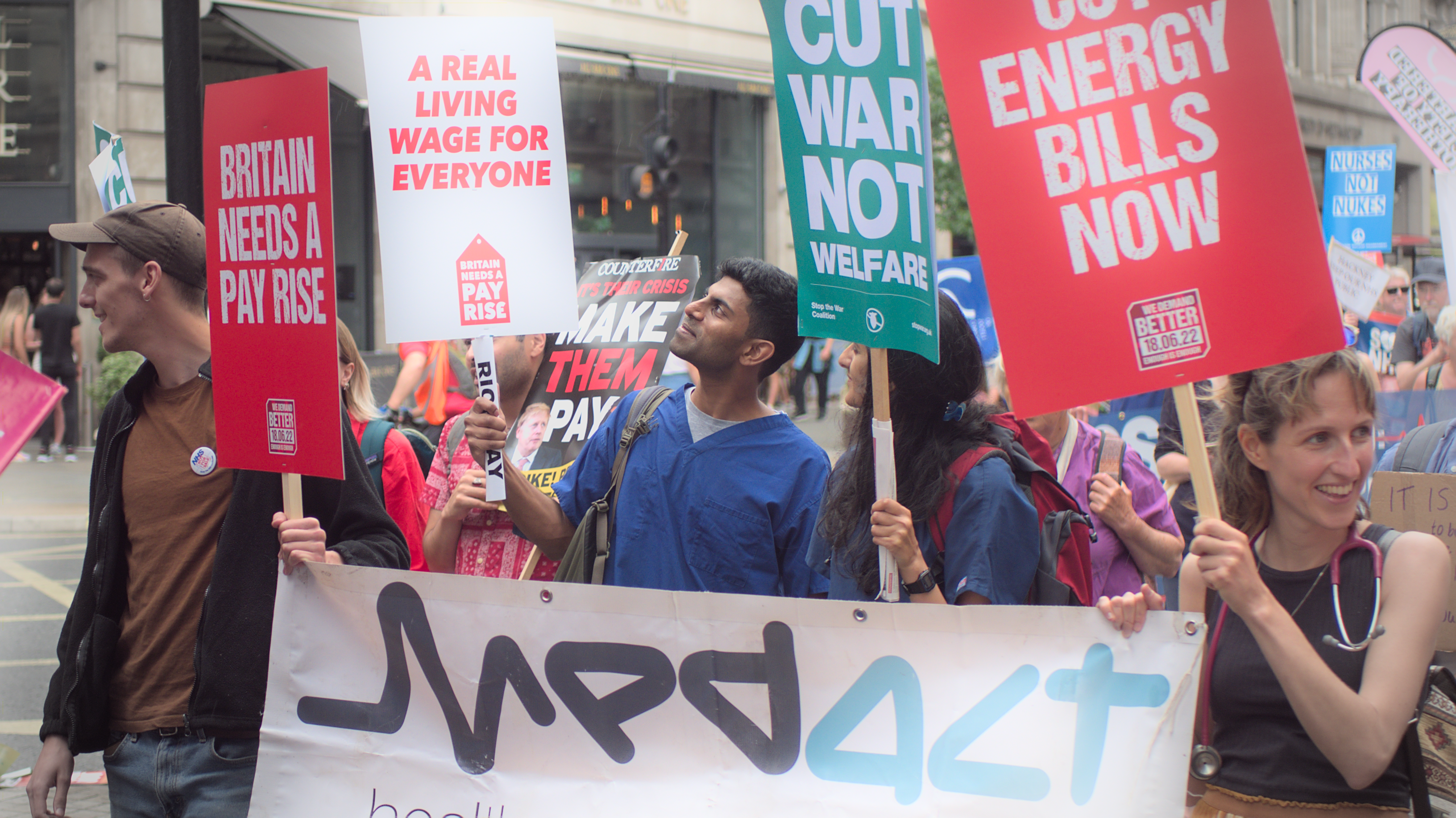 Health workers marching behind a Medact banner. One is wearing scrubs. They are holding placards including 'Britain needs a pay rise', 'Cut war not welfare', 'Cut energy bills now', and 'A real living wage for everyone'.