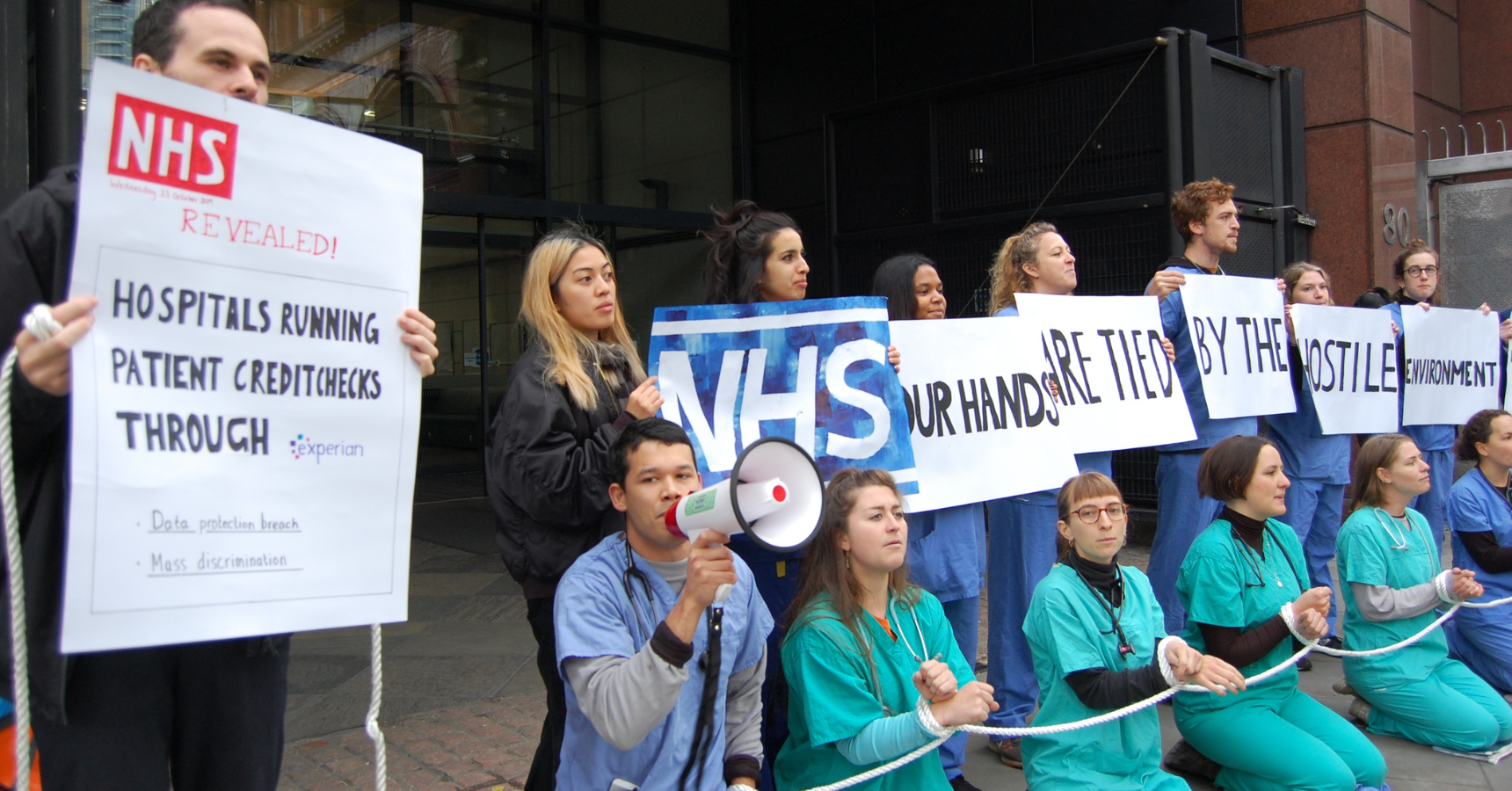 Protestors wearing scrubs in front of a hospital, kneeling with their hands tied together with one length of rope - protestors behind hold signs that together read 'NHS our hands are tied. One placard at the front is a mock newspaper headline reading 'Hospitals running patient credit checks through experian