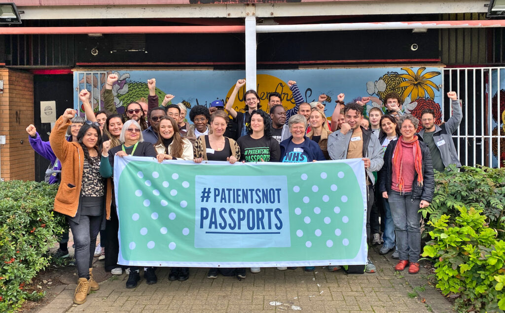 Patients Not Passports campaigners posing for a photo in front of a community centre, holding a large banner with the #PatientsNotPassports logo