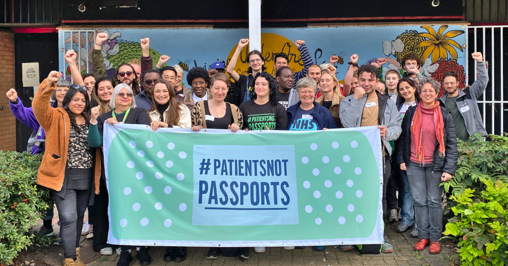 Patients Not Passports campaigners posing for a photo in front of a community centre, holding a large banner with the #PatientsNotPassports logo