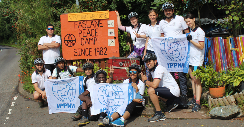 People in bike helmets and all in white t-shirts holding IPPNW flags standing outside in front of an orange sign that says "Visitors welcome! Faslane Peace Camp, since 1982"