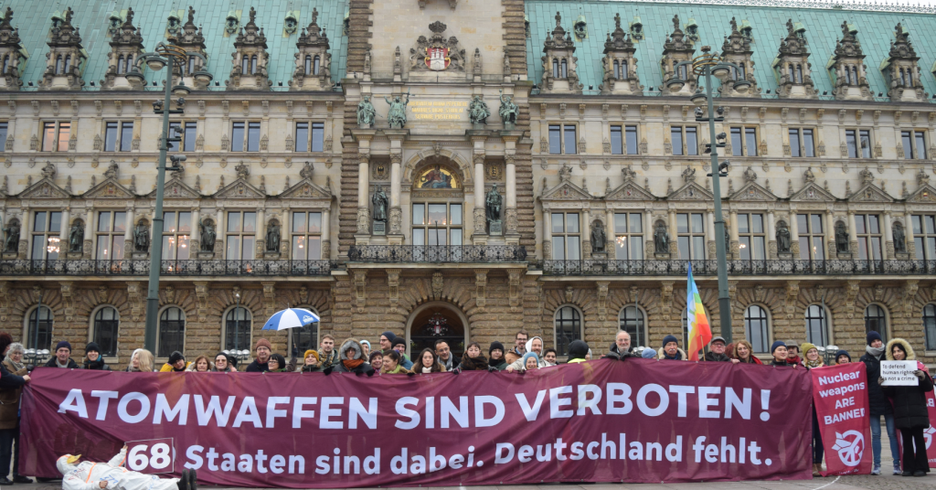 People in warm clothing standing in front of a fancy looking municipal building holding a giant burgundy banner that reads: “Atomwaffen sind verboten! 68 staten sind dabei. Deutschland fehlt.”