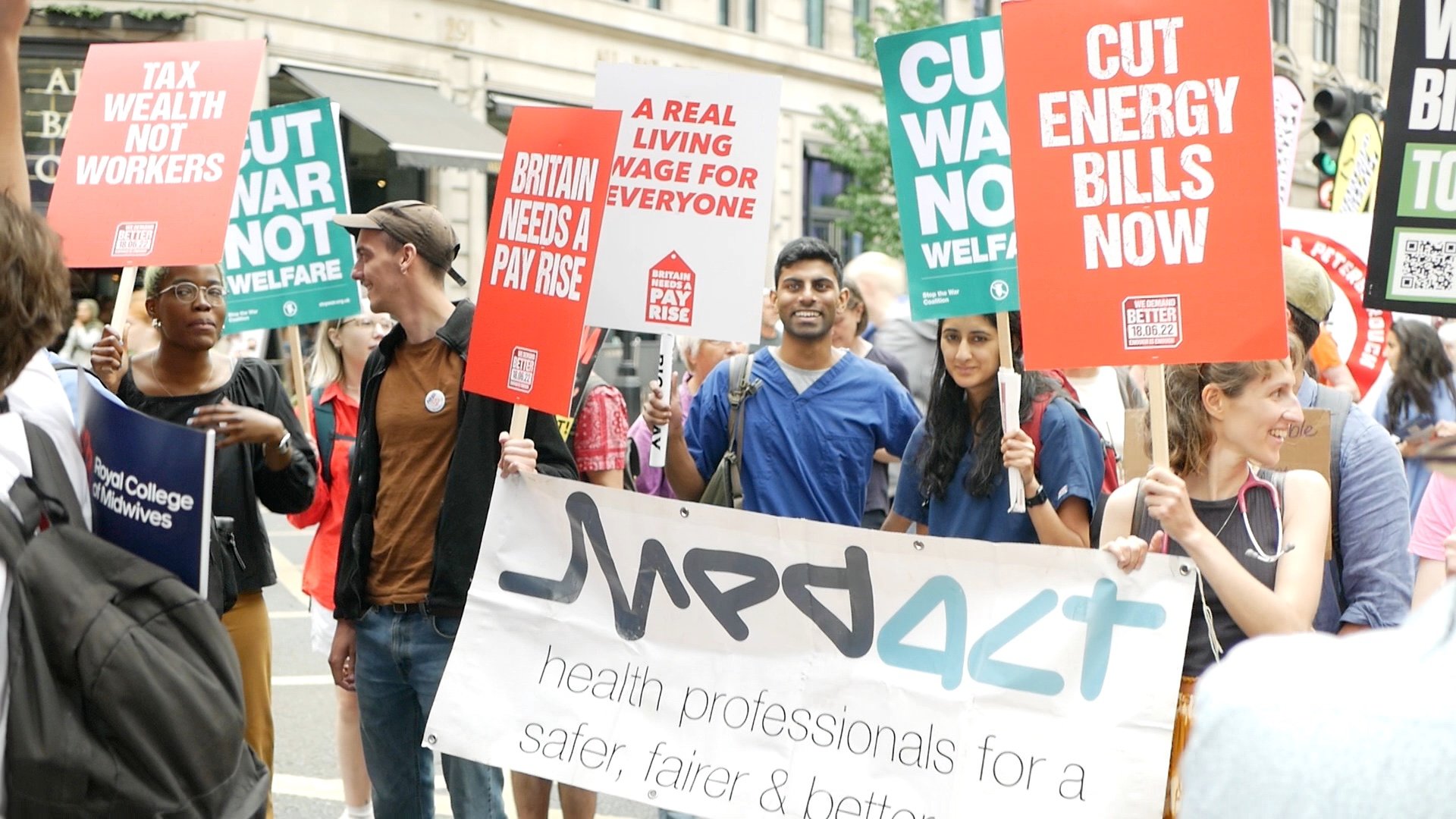 Health workers marching behind a Medact banner. One is wearing scrubs. They are holding placards including 'Britain needs a pay rise', 'Cut war not welfare', 'Cut energy bills now', and 'A real living wage for everyone'.