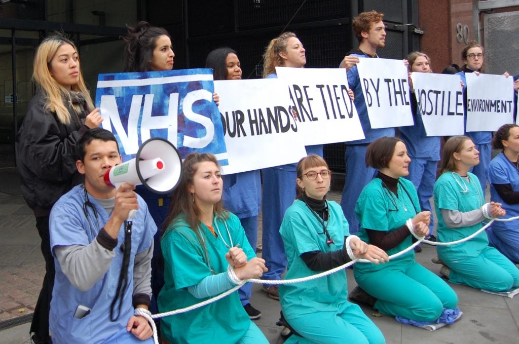 Hospitals running patient credit checks through Experian demonstration. NHS workers with placards and kneeling with hands tied with a rope.