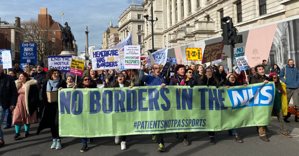 A big crowd of people marching behind a large green banner that reads 'No borders in the NHS #PatientsNotPassports'