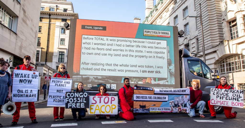 Climate activists in red boilersuits in London outside the Africa Energies Summit in front of a huge TV on a van. The TV shows a slide that reads: 'Frank, Uganda – "Before TOTAL. It was promising because I could get what I wanted and I had a better life this was because I had no fears and worries in this state. I was capable to own and use my land and the property on it freely. After realising that the whole land was taken, I was choked and the result was pressure. I am ever sick."
The activists hold banners and signs such as "End Fossil Fuel Colonialism" and "East Africa Crude Oil Pipeline = Climate Bomb"