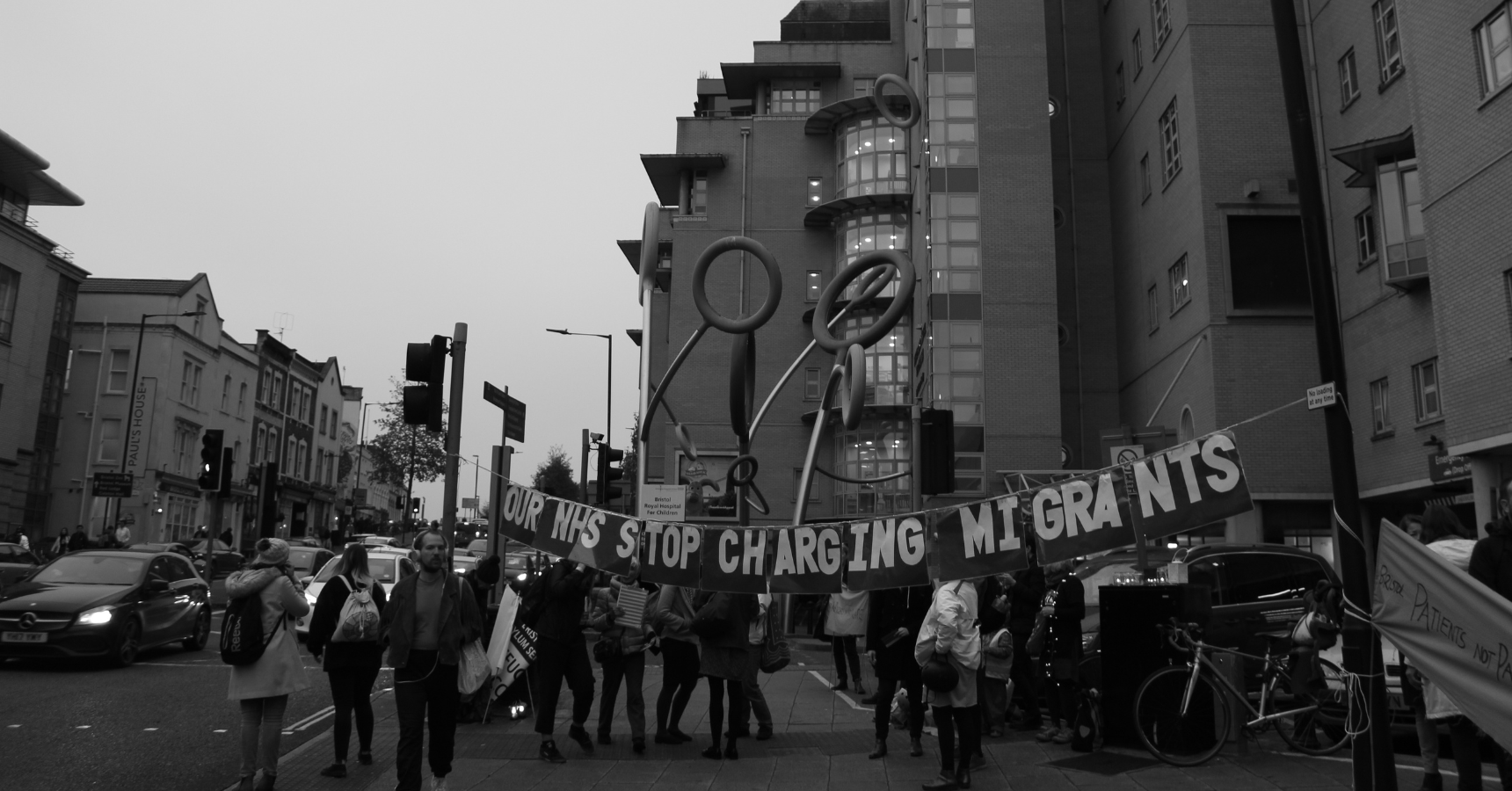 A black and white image of a street protest, with people standing around bunting hanging from a lamp post and traffic light post, that reads: "Our NHS, stop charging migrants"