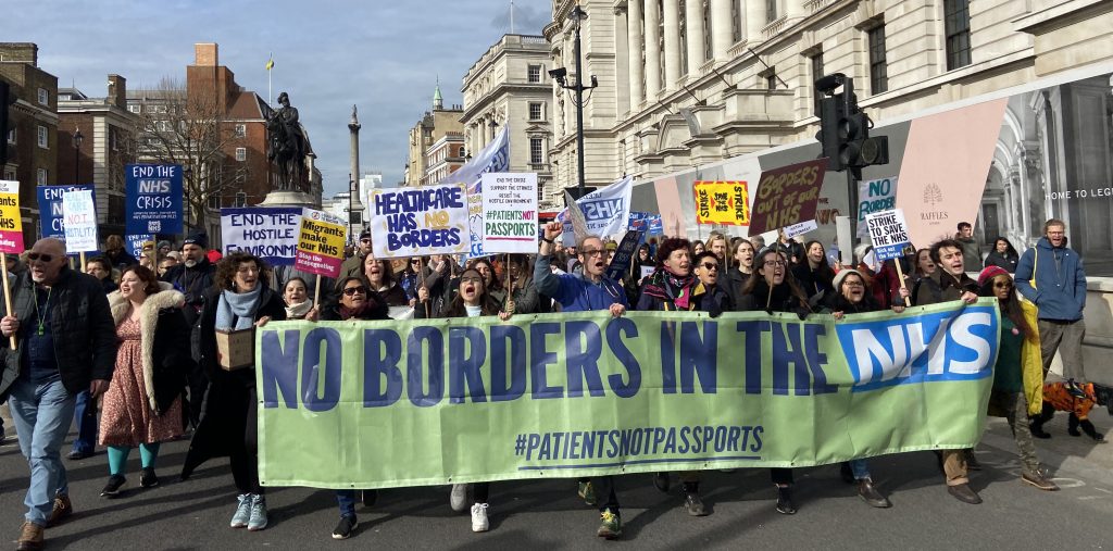 A photo of many people marching in central London behind a huge green banner: ‘No Borders in the NHS #PatientsNotPassports