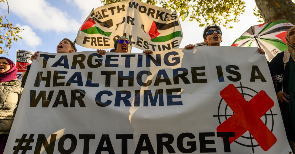 Health workers marching behind a banner that reads 'Targeting healthcare is a war crime #NotATarget'. Another banner is flying in the background that reads 'Health workers for a free Palestine'