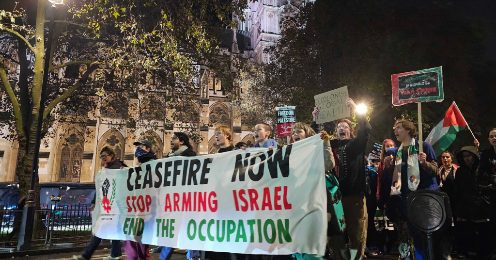 Health workers marching outside Parliament at night behind a banner that reads "Ceasefire now; Stop Arming Israel; End the Occupation"
