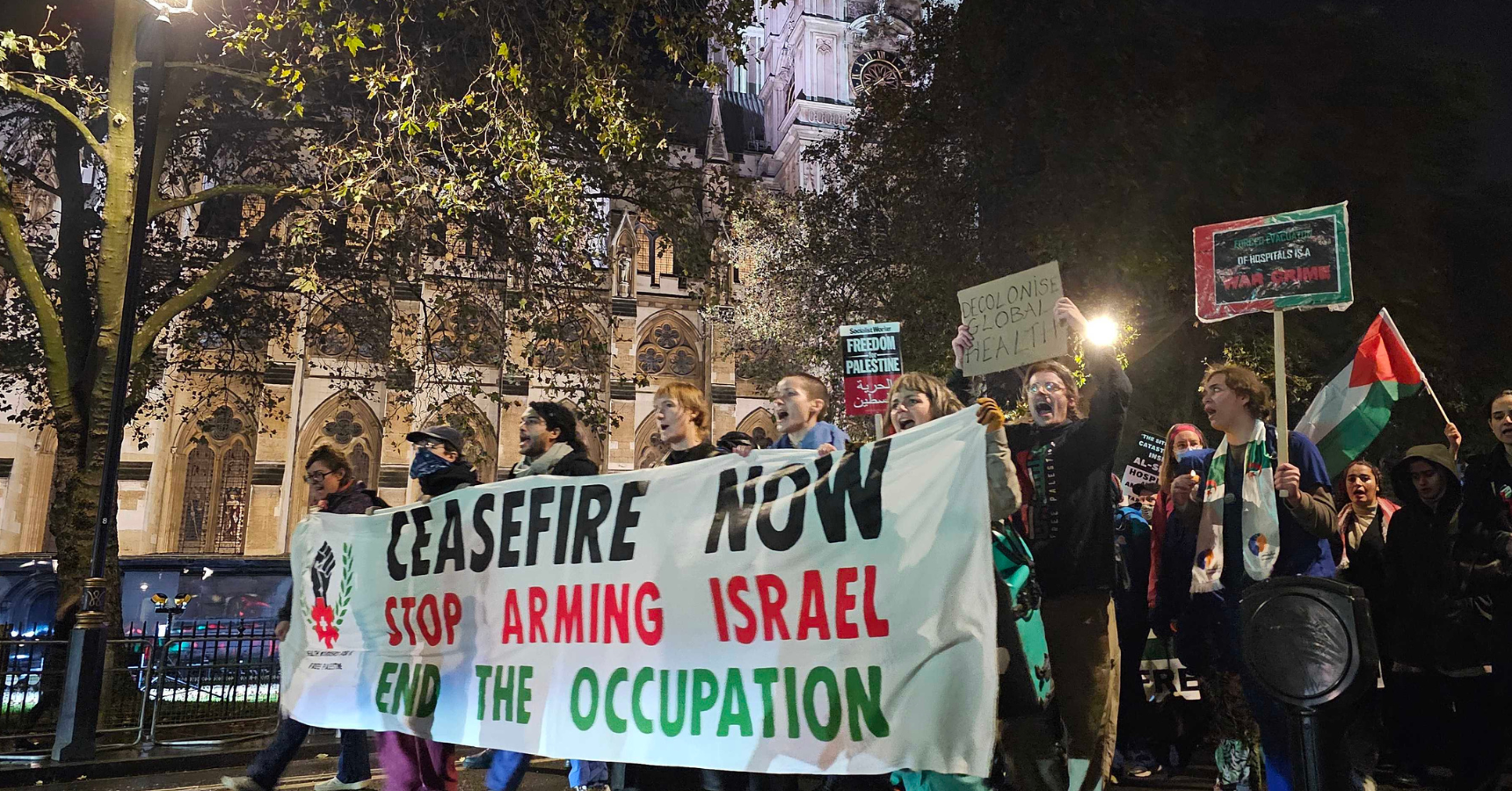 Health workers marching outside Parliament at night behind a banner that reads "Ceasefire now; Stop Arming Israel; End the Occupation"