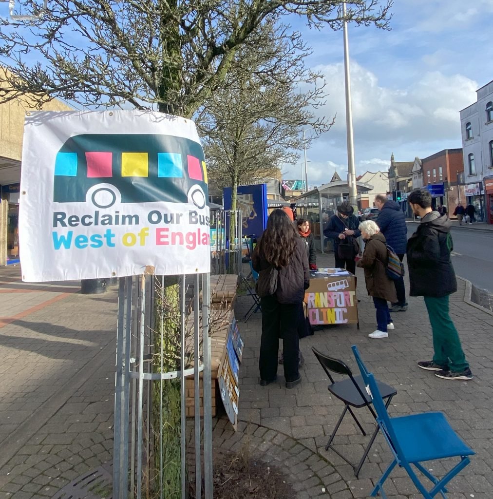 In the background people gather around the transport clinic stall. In the foreground, a poster taped to a tree reads "Reclaim our Buses West of England"