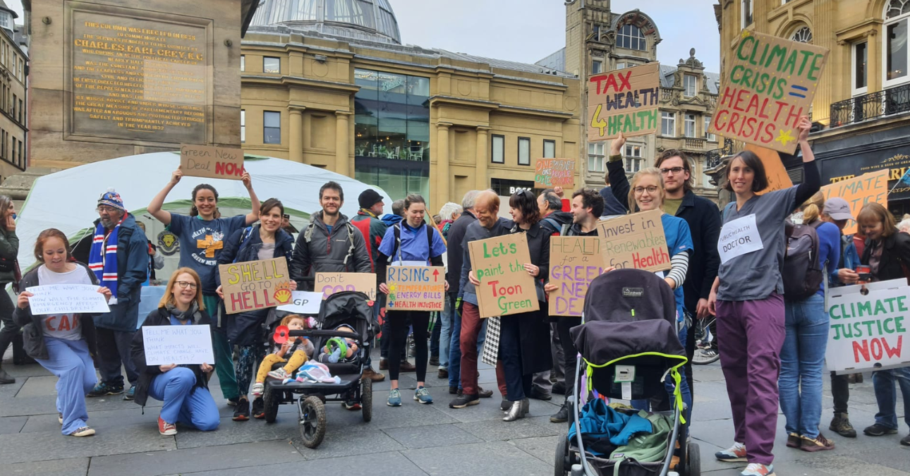 A group of people at a climate protest in the centre of Newcastle, including adults with prams and children, smiling at the camera and holding handmade placards including "Climate Crisis = Health Crisis" and "Let's paint the Toon Green".