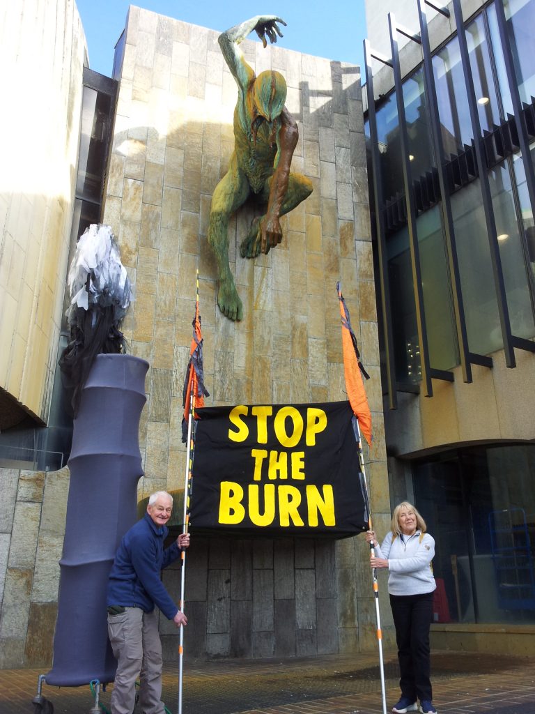 Two protestors with a large chimney model, holding a large banner with yellow text on black background reading "Stop the Burn"