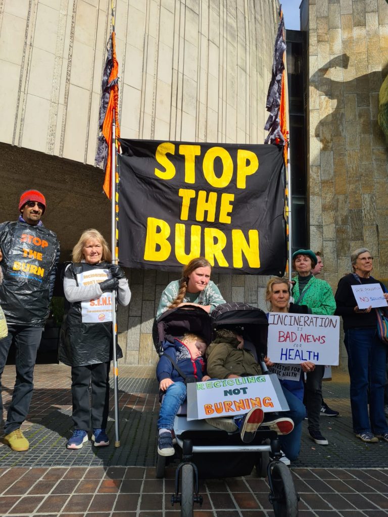 Protestors including two children in one pram. The children have a placard that reads "Recycling Not Burning". A protestor in scrubs to their right has a sign that reads "Incineration is bad news for health". They are all in front of a large banner that reads "Stop the Burn"