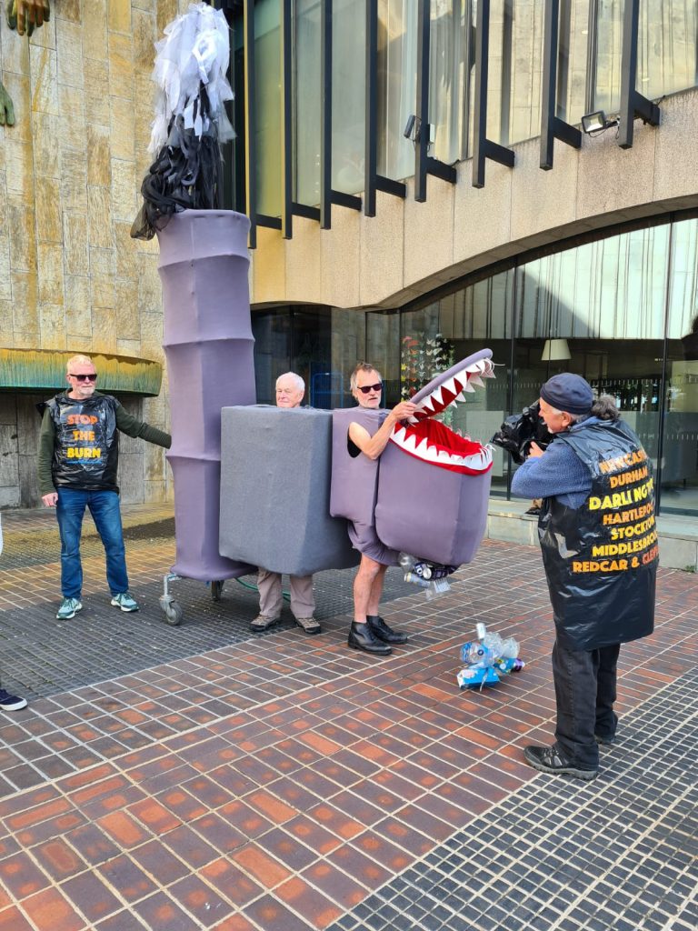 Protestors wearing a large model incinerator costume, with a large mouth and sharp teeth, while another protestor dressed in bin bags throws rubbish into the mouth