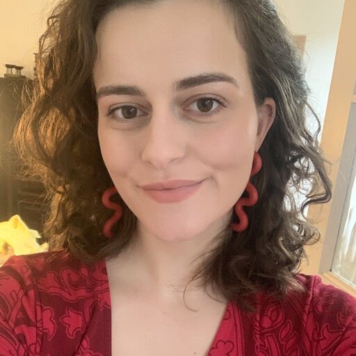 A fair-skinned woman smiling, wearing a red top and long, red, wavy earring