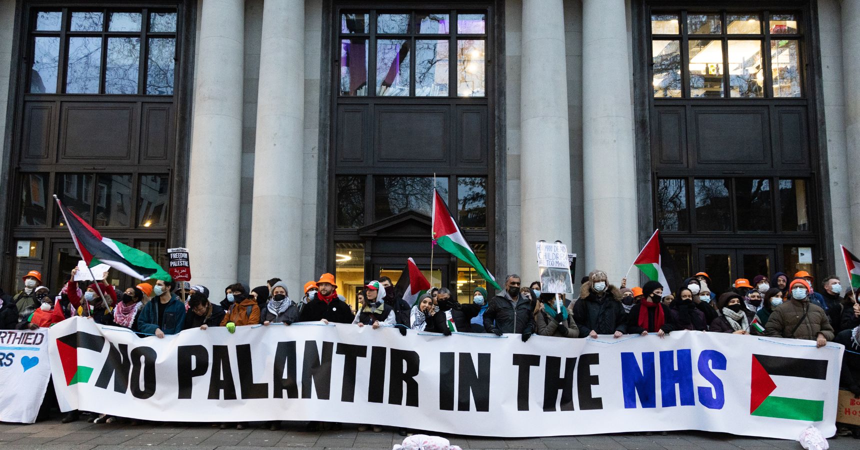 Protesters in front of Palantir's London HQ behind a No Palantir in the NHS banner with Palestine flags.