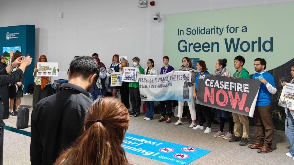 People lined up in a protest, with banners including 'Ceasfire now', and 'No Nukes, No War, No Warming'.