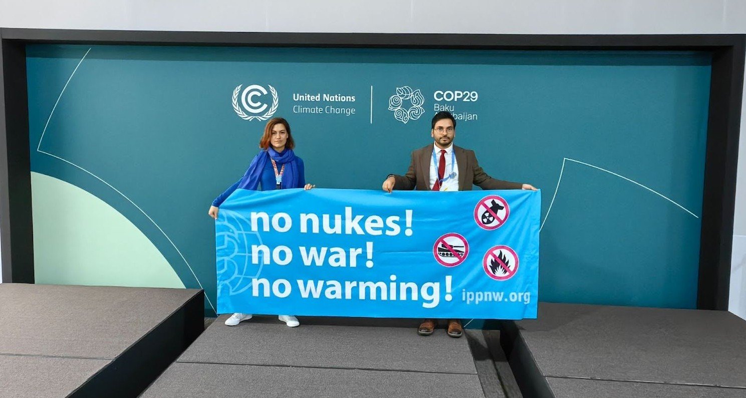 No Nukes! No War! No Warming! - Banner being held by two people in from of a COP29 backdrop