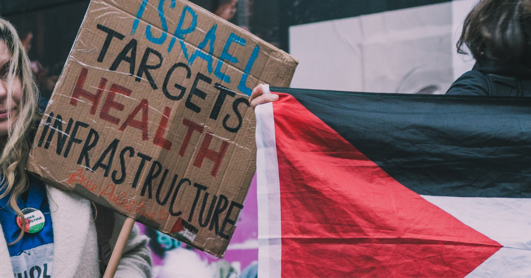 A placard at a demonstration next to a Palestinian flag. Placard reads: Israel targets health infrastructure