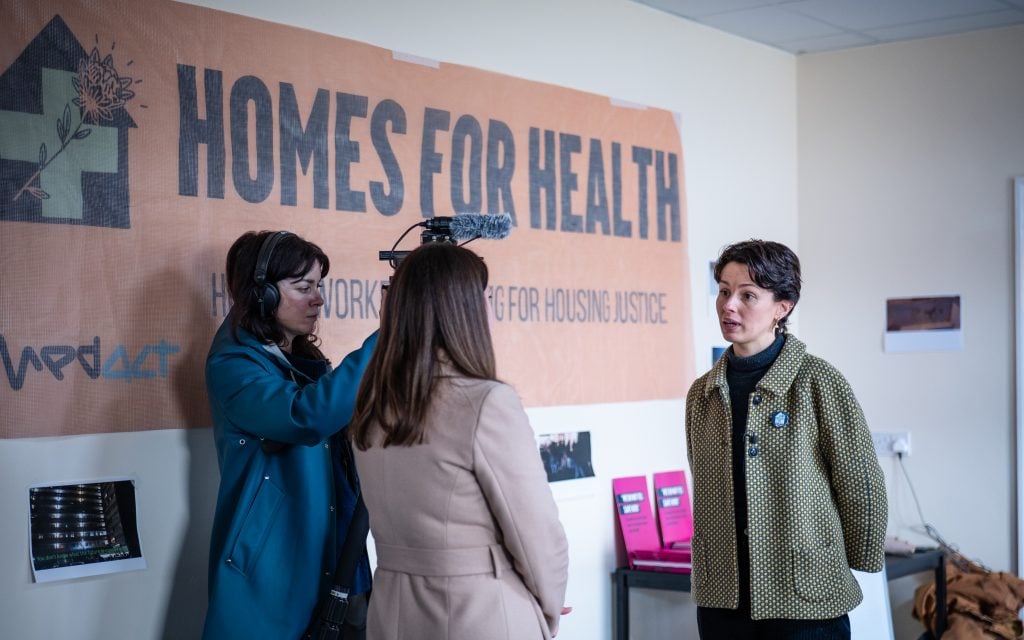 Medact Bristol member Amelia doing an interview in to a camera and presenter, standing in front of a large orange 'Homes for Health' banner