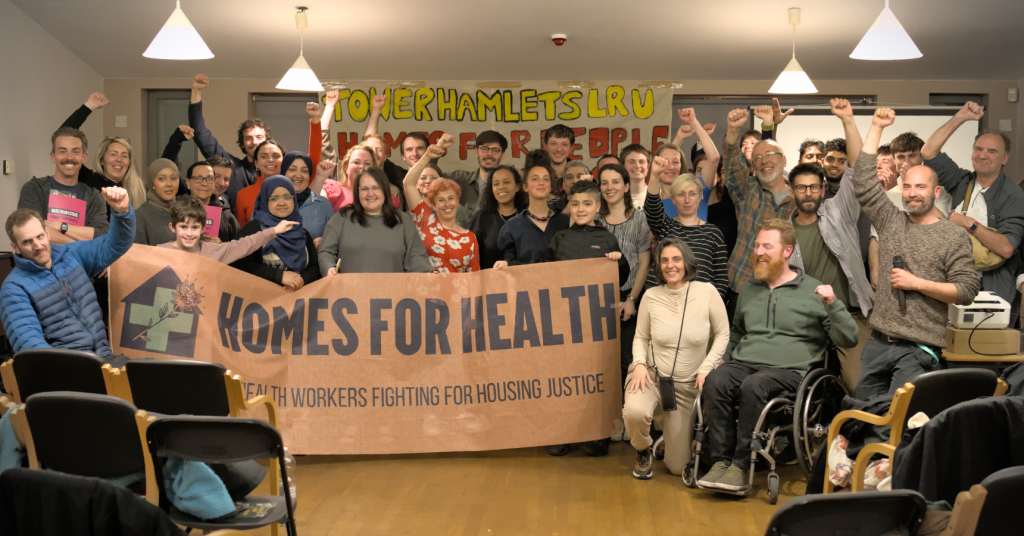 A crowd of people posing for the camera in a room with fists raised in the air, gathered around a large 'Homes for Health' banner