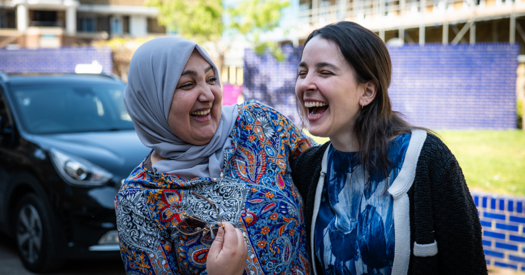 A white woman and a brown hijabi woman laughing together in a car park