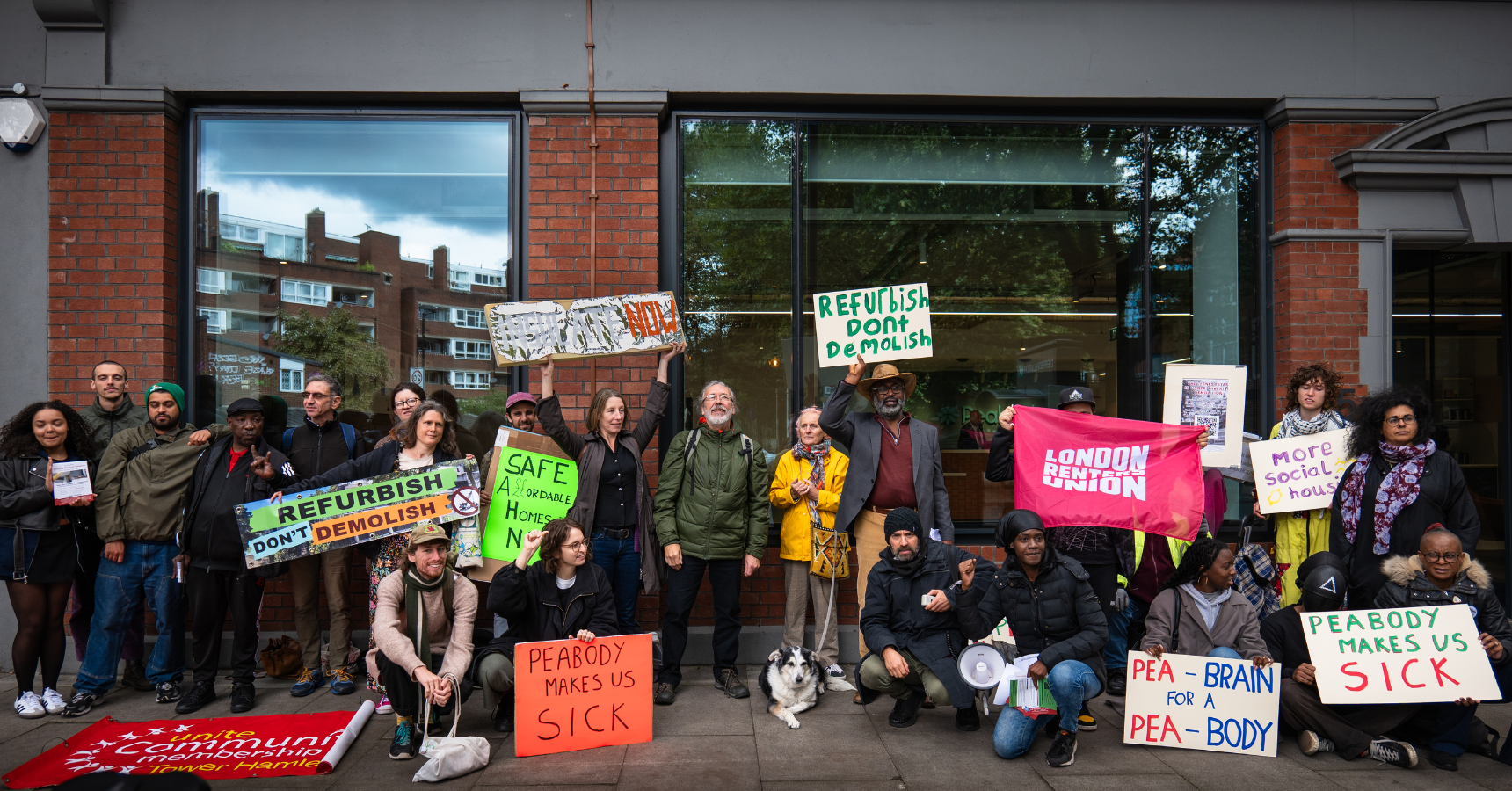 A crowd of protestors lined up outside a glass-fronted building holding housing justice placards and smiling at the camera