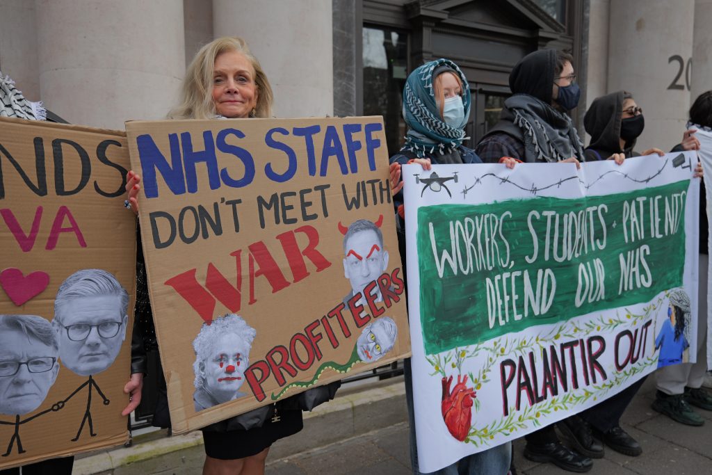 A protestor with blonde hair looking at the camera, holding a placard that reads 'NHS staff don't meet with war profiteers'.