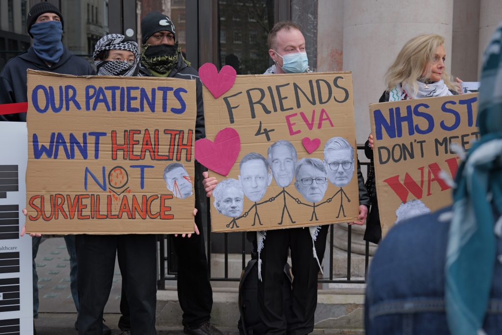Protestors standing in a line with placards that read 'Our patients want health not surveillance', and 'Friends 4 eva' (images of Palantir leaders and Jeffrey Epstein).