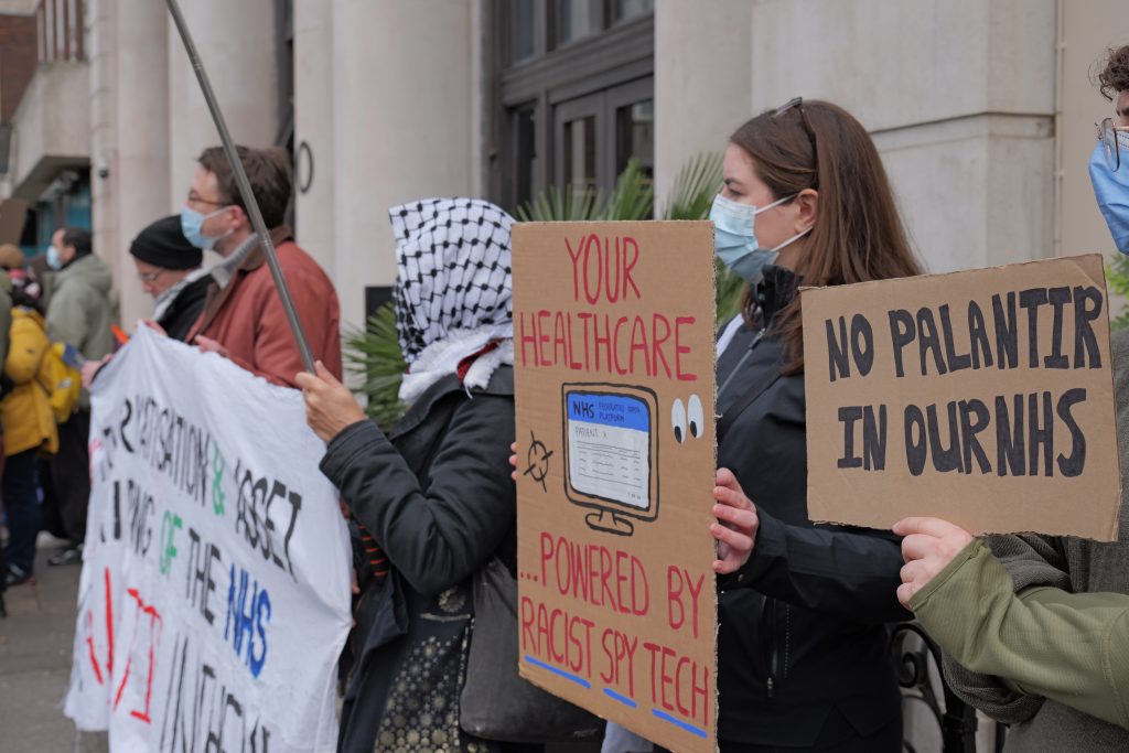 Protestors, with one holding a placard that reads 'Your healthcare, sponsored by racist spy tech', and another holding one that reads 'No Palantir in the NHS'