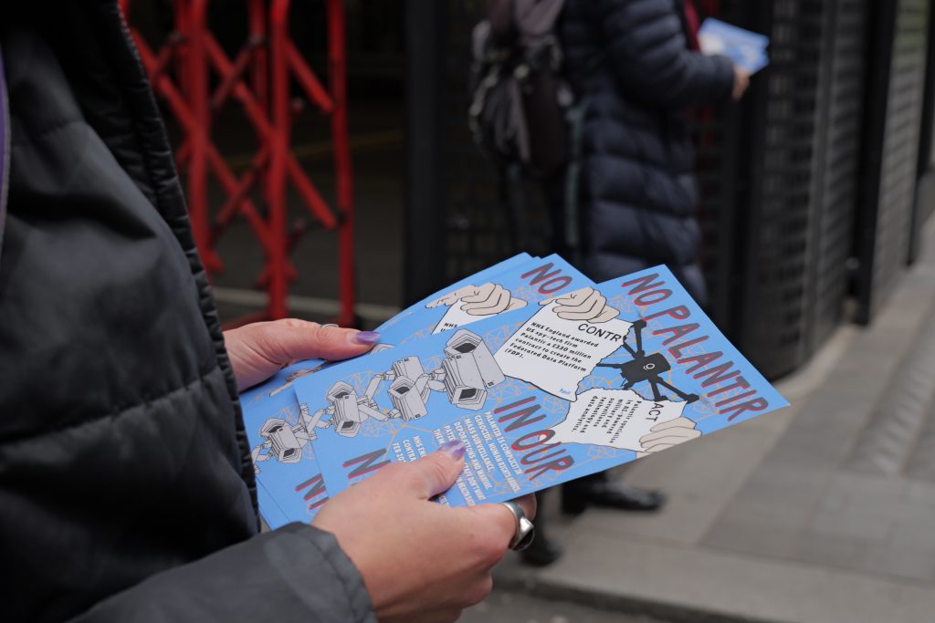 A photos of someone's hands holding flyers in a street – the flyers reads 'No Palantir in our NHS' and has an image of a drone