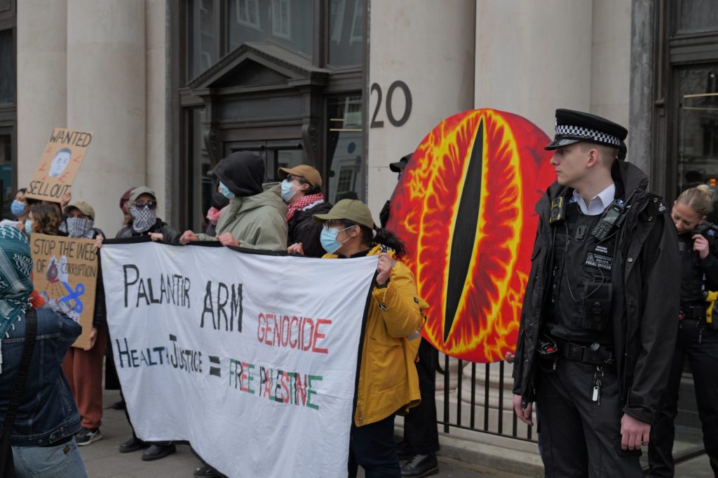 Protestors with a policeman standing next to them. The protestors hold up a giant eye of Sauron, and a banner reading 'Palantir Arm Genocide; Health justice = Free Palestine'.
