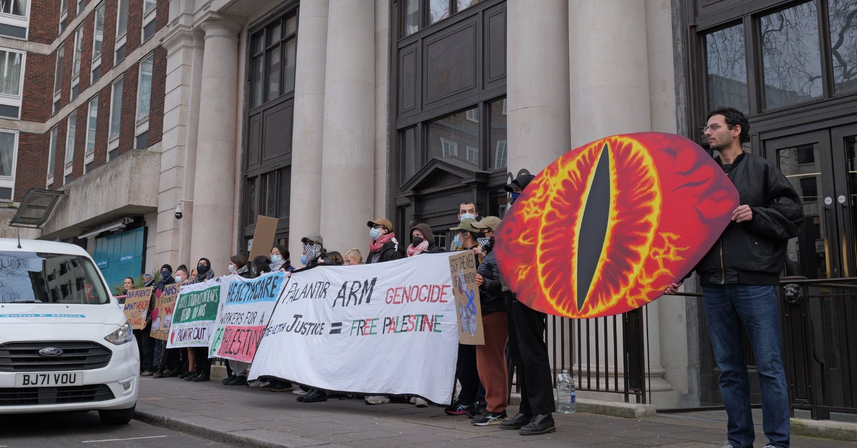 The protest outside Palantir's London HQ, where a group of people stand in a line, holding banners, placards, and a giant eye of Sauron model